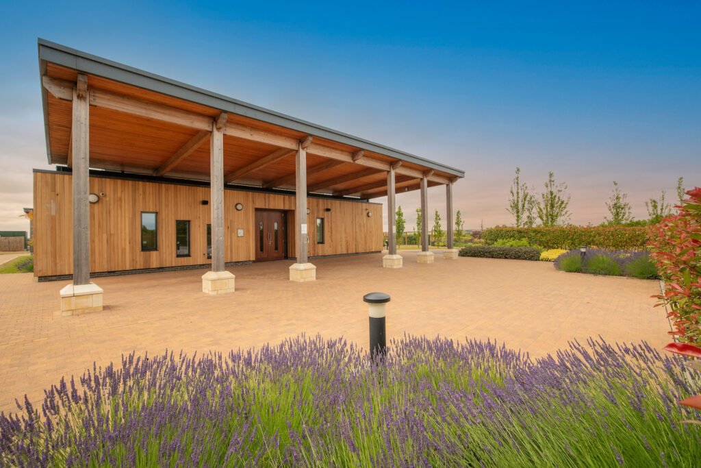 Outside of Cam Valley Crematorium showing a rectangular building covered in wooden slats with an overhang and paved driveway surrounded by lavendar