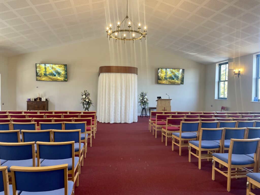 A large chapel with white walls and red carpet with chairs either side of a wide aisle leading to a catafalque with closed curtains and a lectern