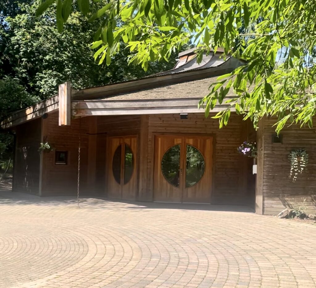 Exterior of Greenacres Epping Forest Park Woodland Hall - a wooden octaganol building sitting on the edge of a circular paved courtyard surrounded by trees