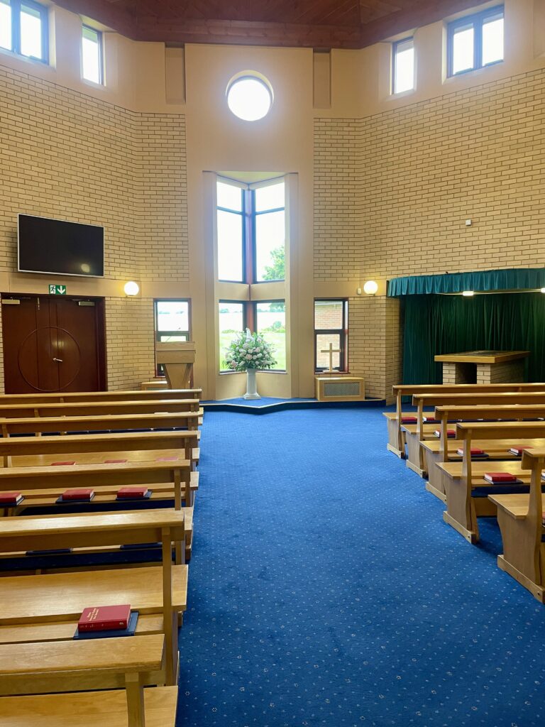 A chapel with pale brivh walls, blue carpet and wooden pews with a tall narrow window overlooking the countryside