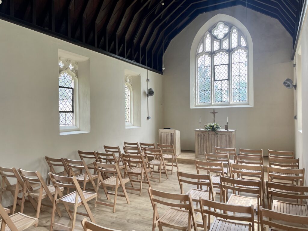 Inside a small chapel with pale stone walls and wooden vaulted ceiling set out with wooden chairs and a small alter and a cross beneath an arched window