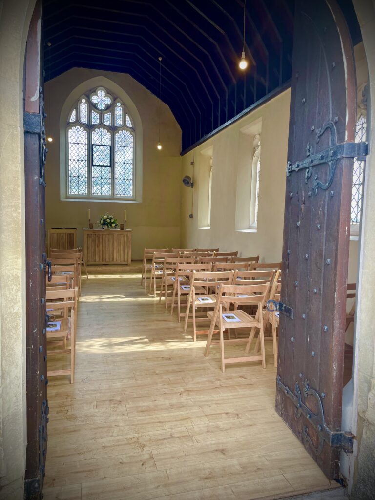 Looking through the open wooden doors into a small light chapel set out with chairs either side of an ailse and an small alter with candles and flowers at the end