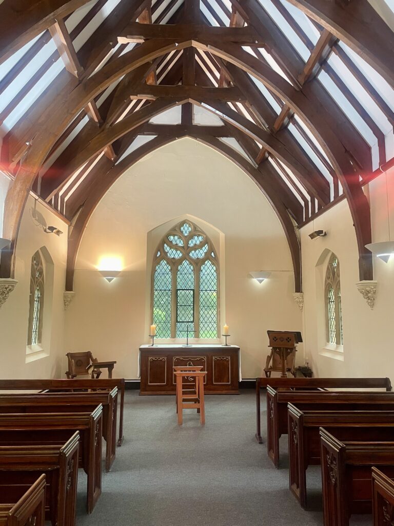 A small chapel with light walls and wooden vaulted ceiling with wooden pews either side of an aisle leading to a lectern and alter