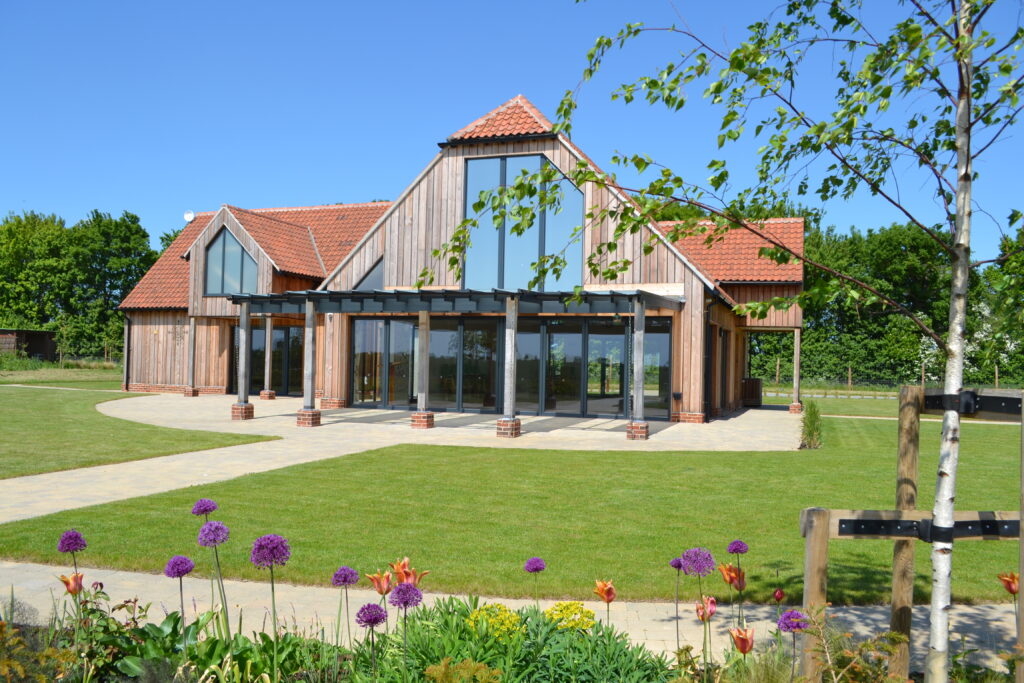 A wooden building with large glass window in the vaulted apex looking over a paved terrace and pathways leading to a lawn and wild flowers