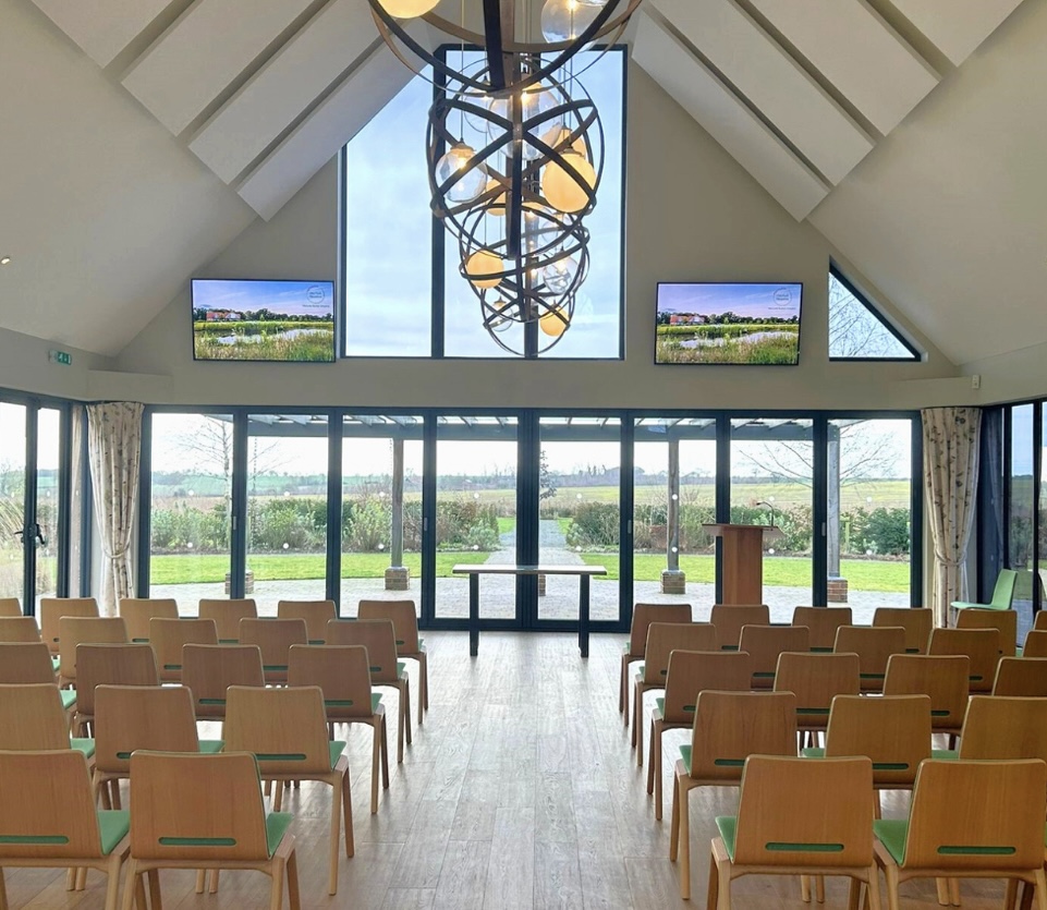 A large and bright room with fully retractable glass bifolding doors at the end looking out to the meadows, with chairs laid out wither side of an aisle