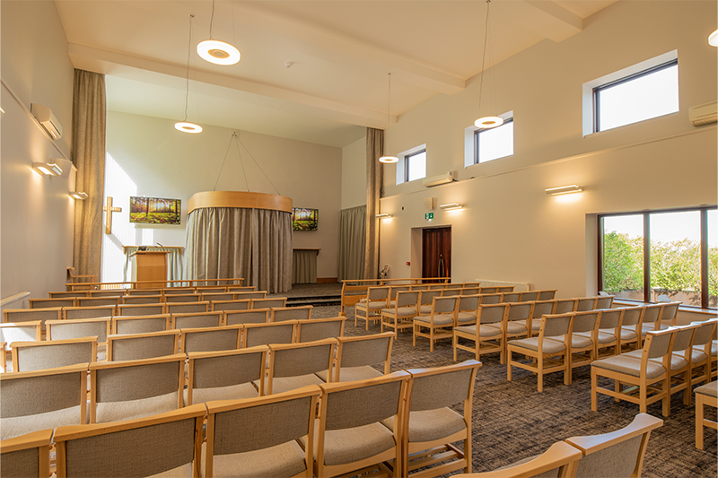 A bright chapel with chairs either side of an aisle leading to a curtain and a lecturn