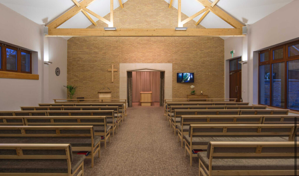 A light chapel with a stone wall opening to a wooden catafalque at the end of an aisle with pew seating on either side